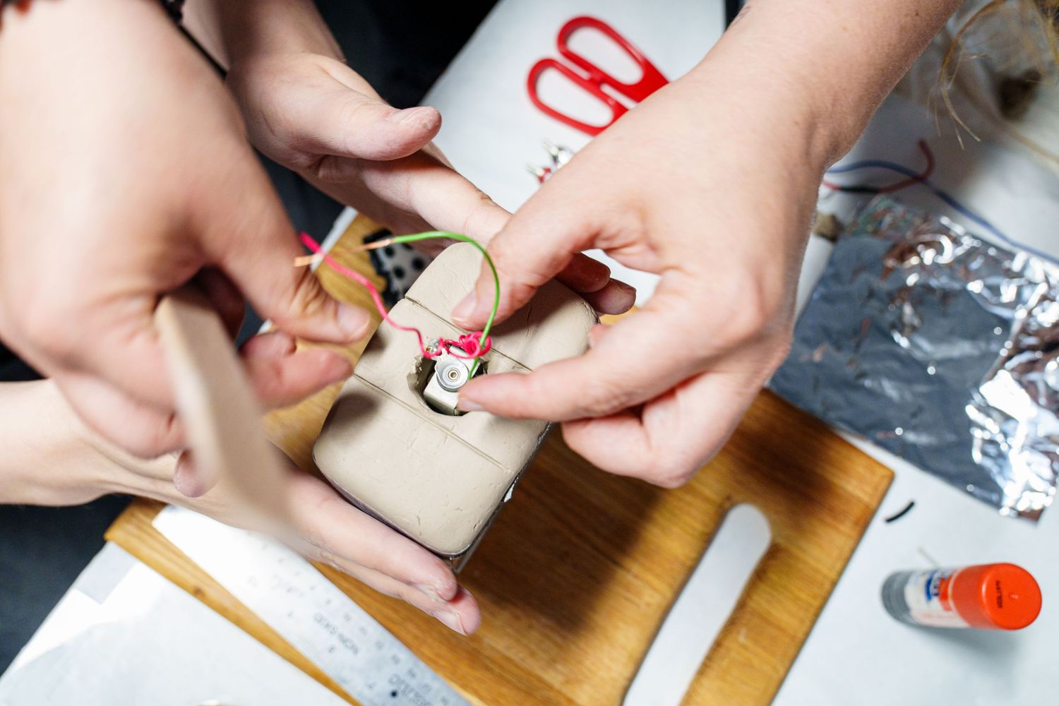 Hands assembling a small electronic circuit with colorful wires on a wooden cutting board in a workshop.