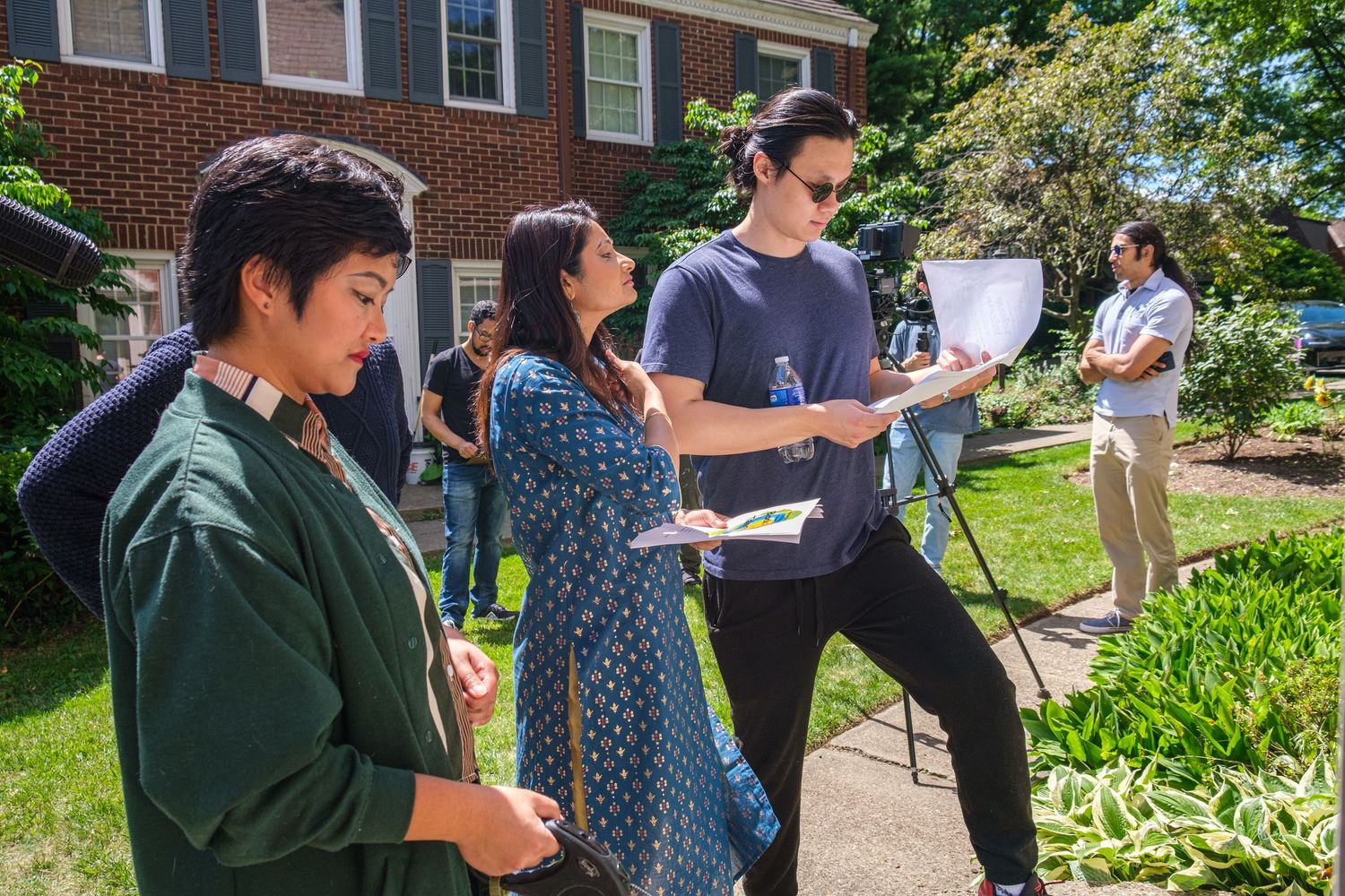 Students gather outdoors on a sunny day, reviewing papers near a brick building with lush green landscaping.