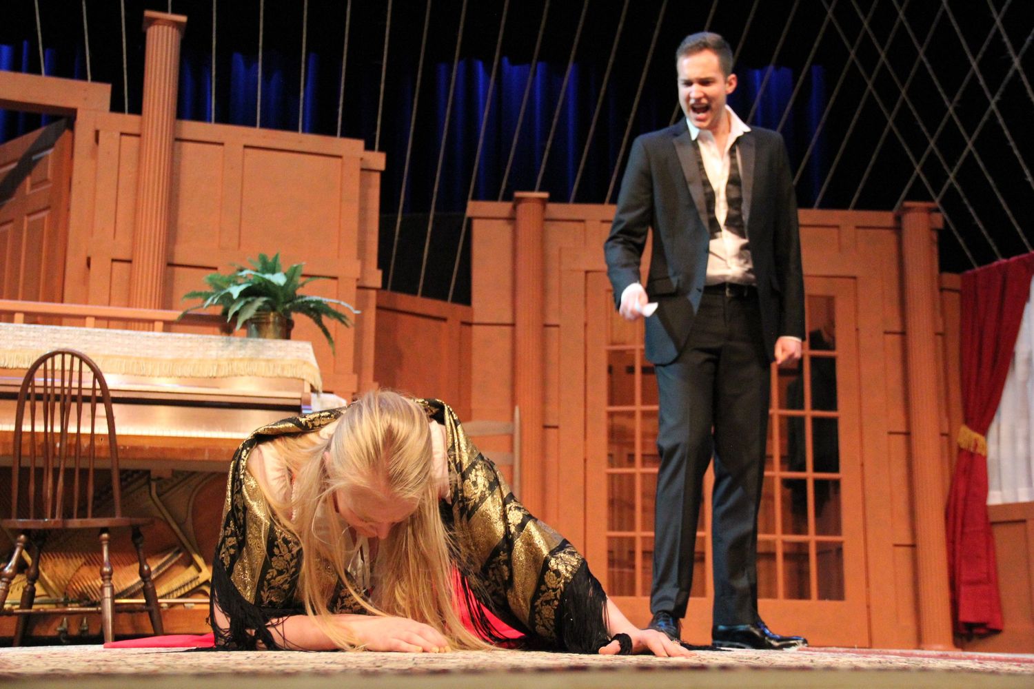 Stage performers act in a dramatic scene on a theater set with red curtains and props.