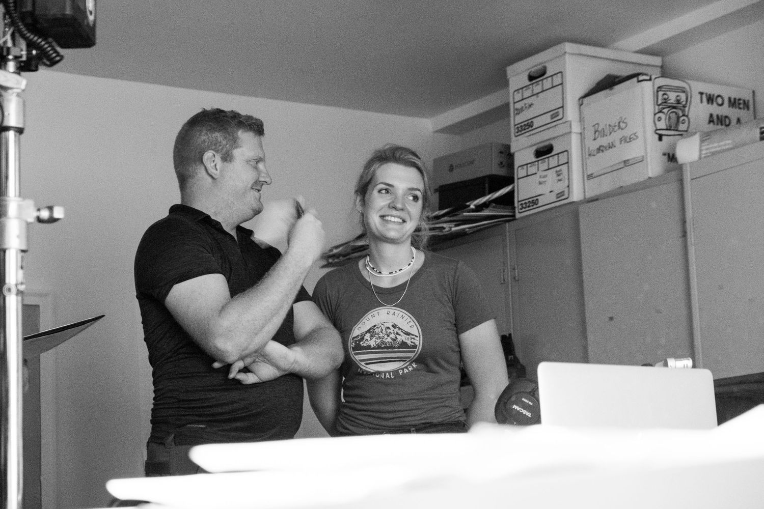 Behind-the-scenes black and white photo of a man applying makeup to a smiling woman on a film set.