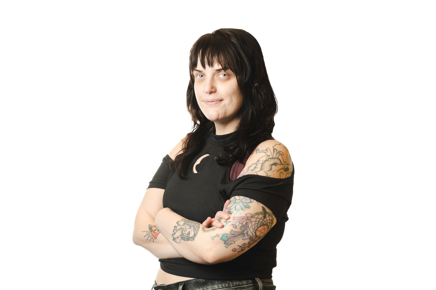Tattooed woman with dark hair standing with arms crossed wearing a black top against white background.