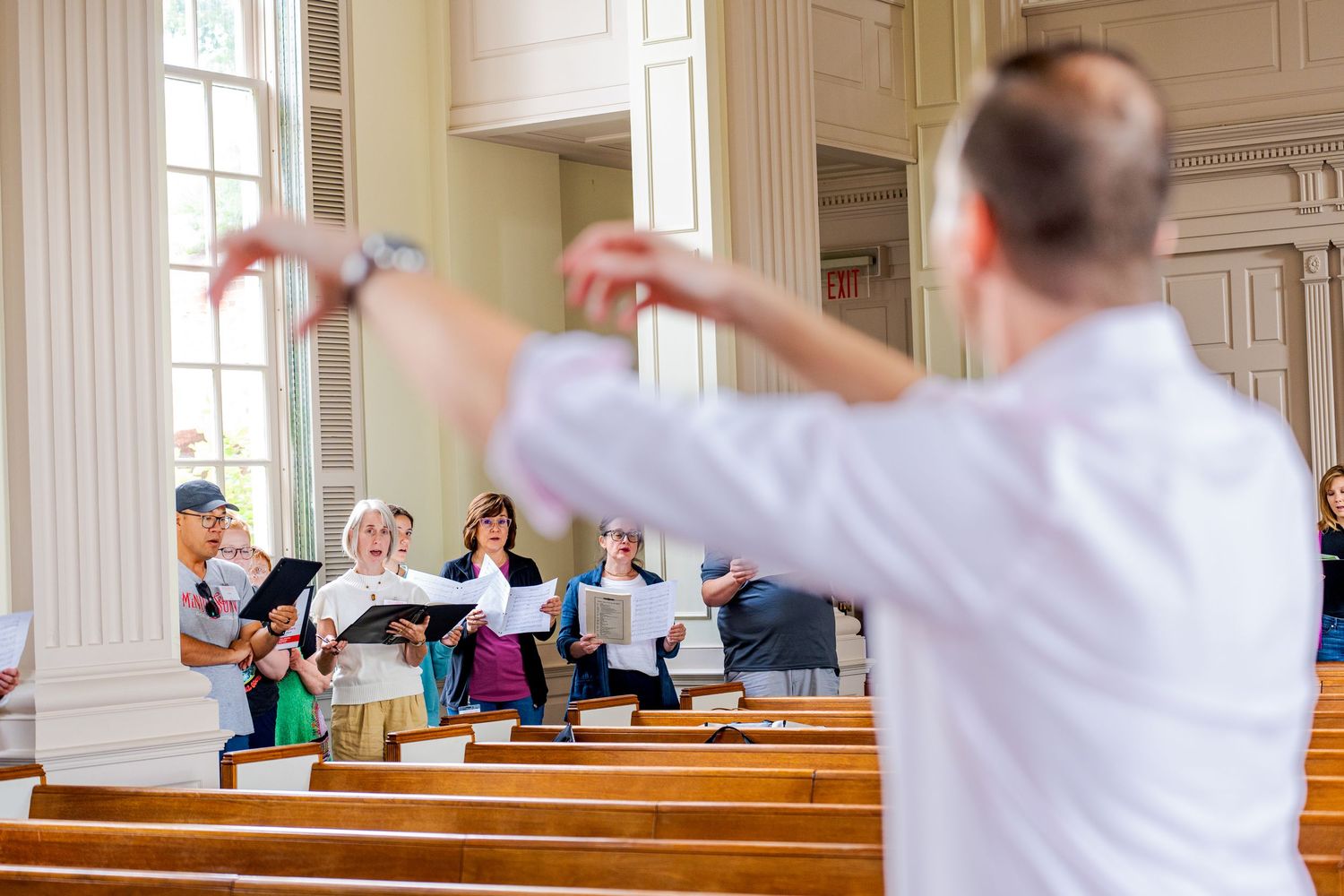 Choir director conducting singers holding sheet music in a classic church interior.