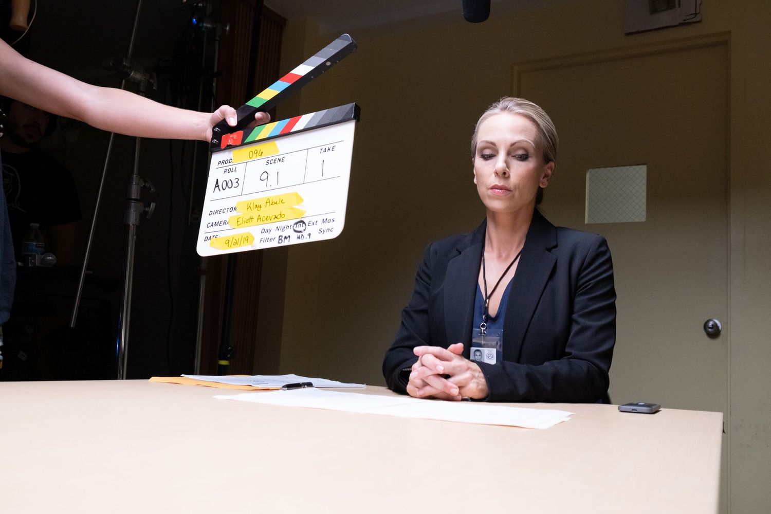 A woman in a black blazer sits at a table as a clapperboard is held above during filming.