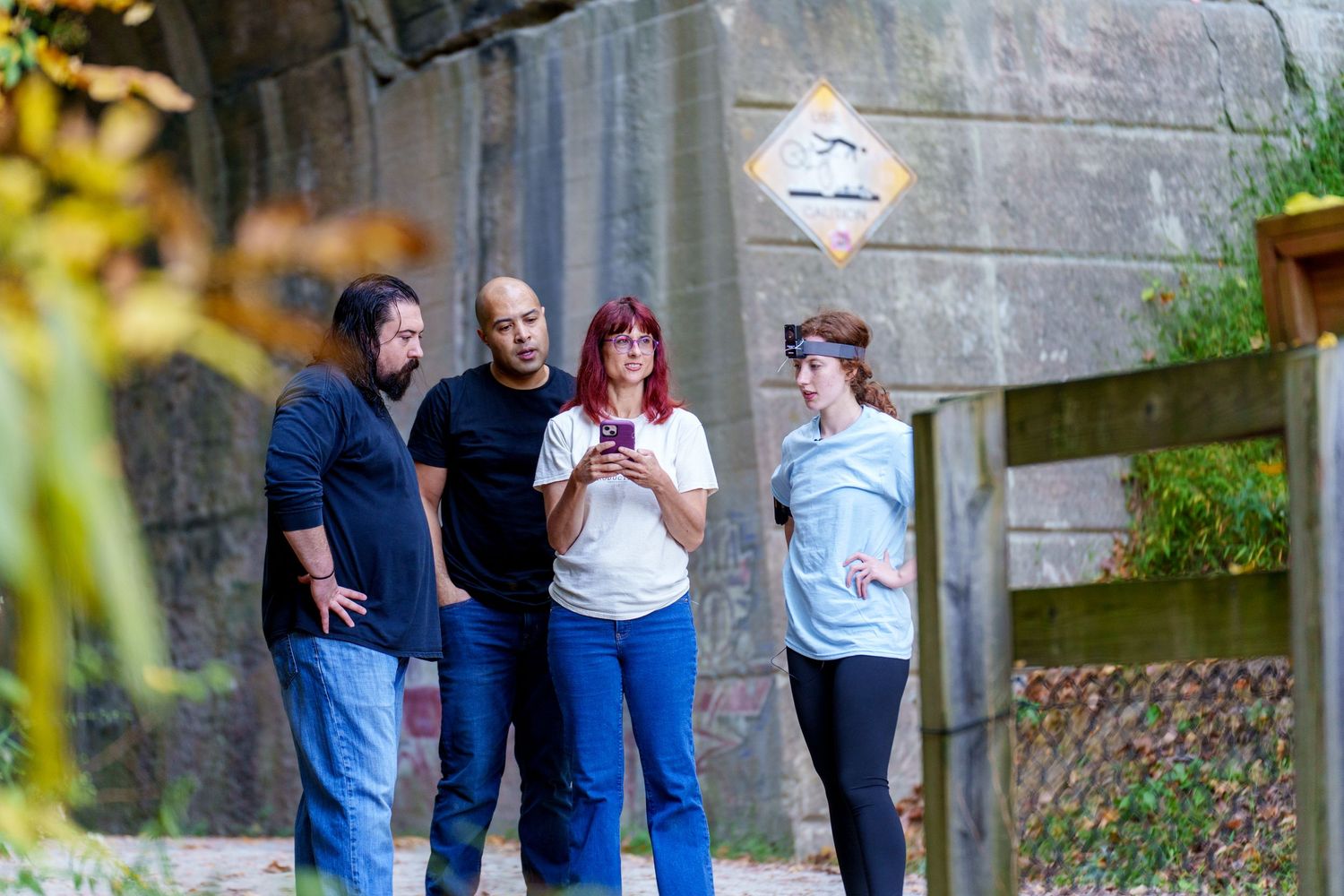 Four people stand outdoors near a stone wall, looking at a smartphone together.