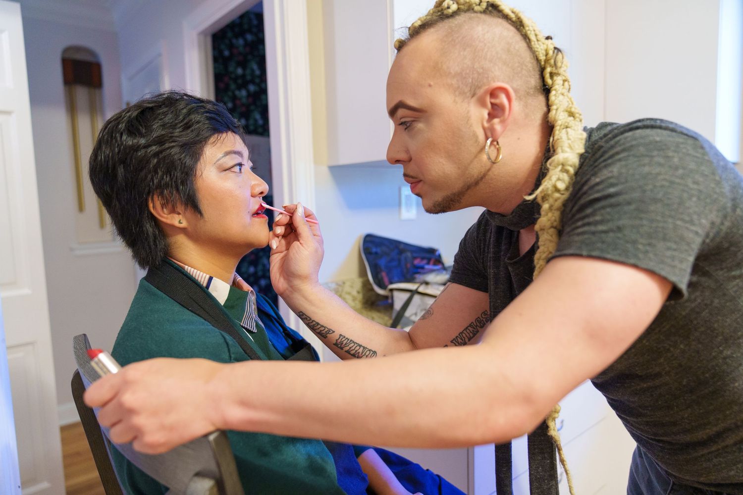 A tattooed makeup artist with a mohawk applies lipstick to a young person sitting in a chair indoors.