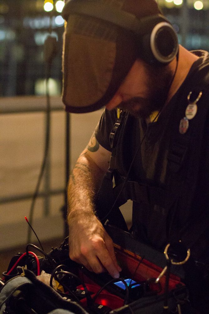 A bearded man wearing goggles and headphones works on electronic equipment with colorful wires at night.