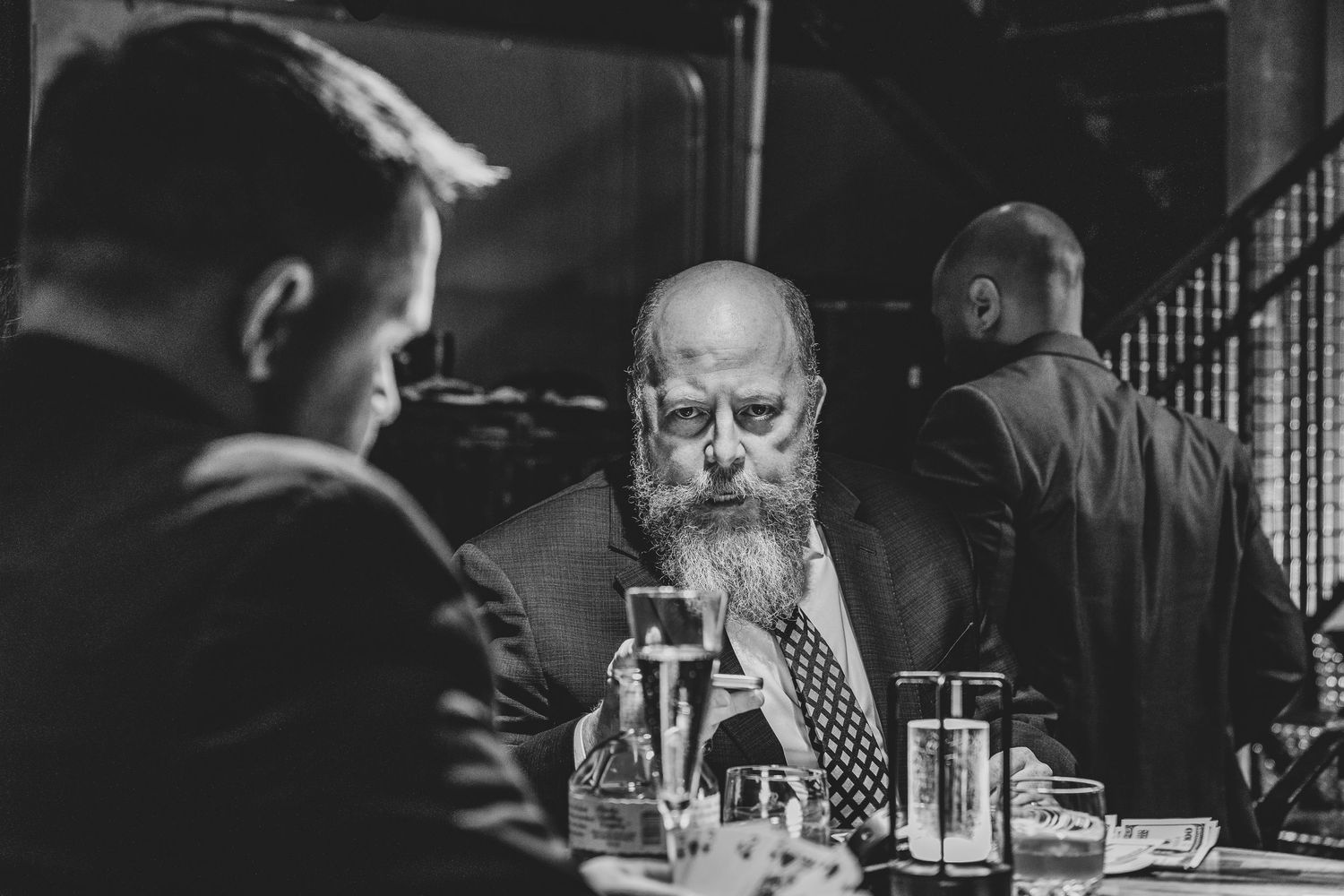 Black and white photo of people socializing at a dimly lit bar with glasses and bottles on tables.