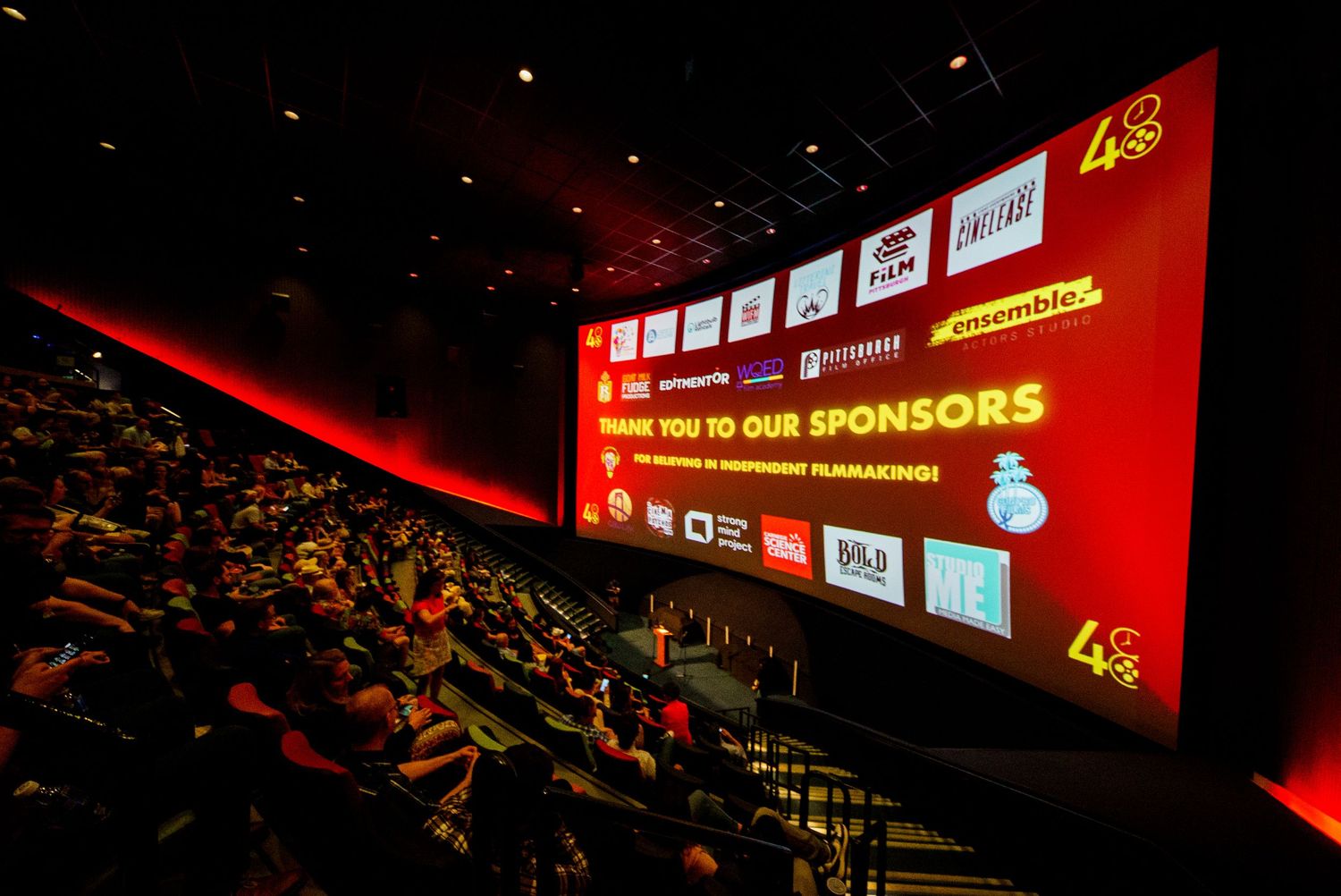 Interior view of a large dark theater auditorium with red lighting and digital displays showing sponsor information.