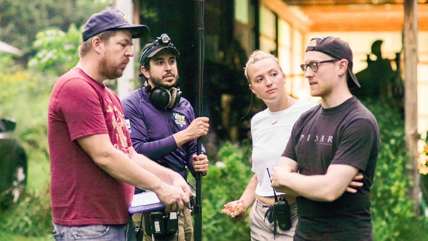 Four male film crew members having a discussion on an outdoor movie set surrounded by greenery.