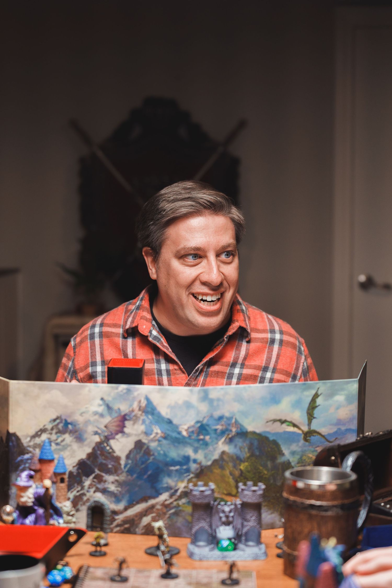 A smiling man in a plaid shirt plays a fantasy tabletop RPG with miniatures and a mountain scene game master screen.