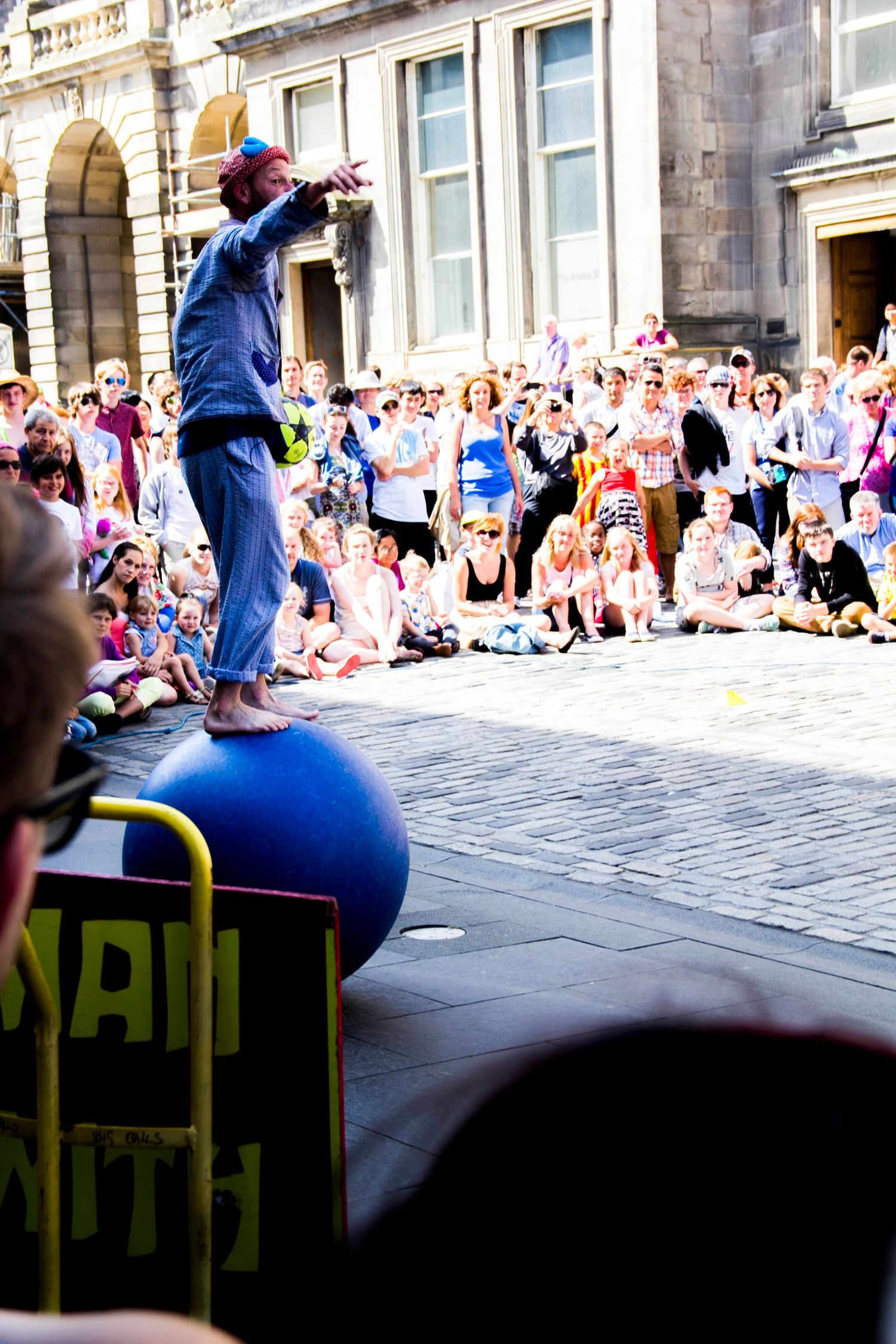 Street performer balances on large ball in front of seated crowd at Edinburgh Fringe Festival.