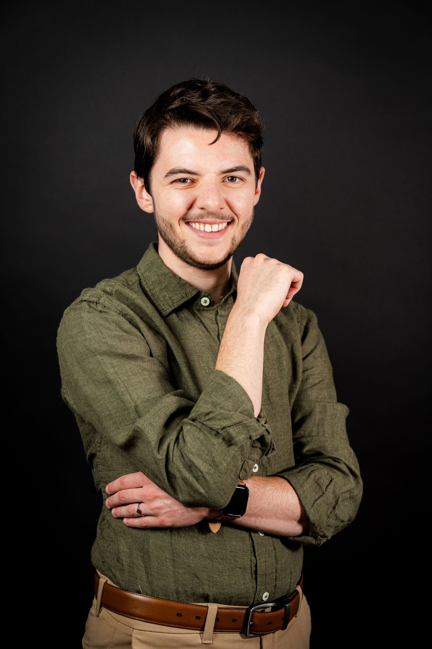 Professional portrait of someone wearing an olive green button-down shirt and khaki pants against a dark background.