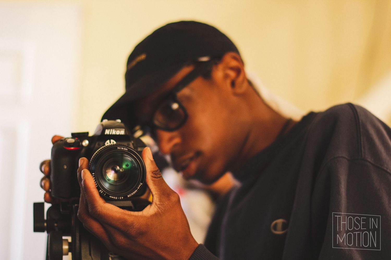 A young man wearing a cap and glasses looks through a Nikon camera on a tripod indoors.