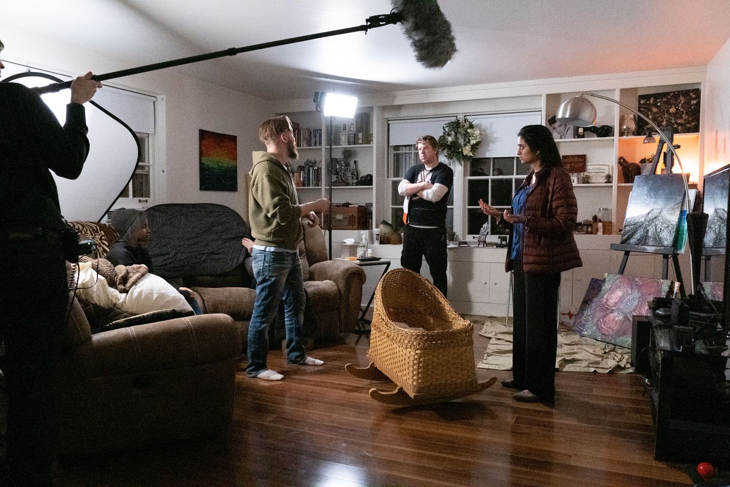 Film crew shooting a scene in a living room with boom mic, lighting equipment, and actors on hardwood floors.