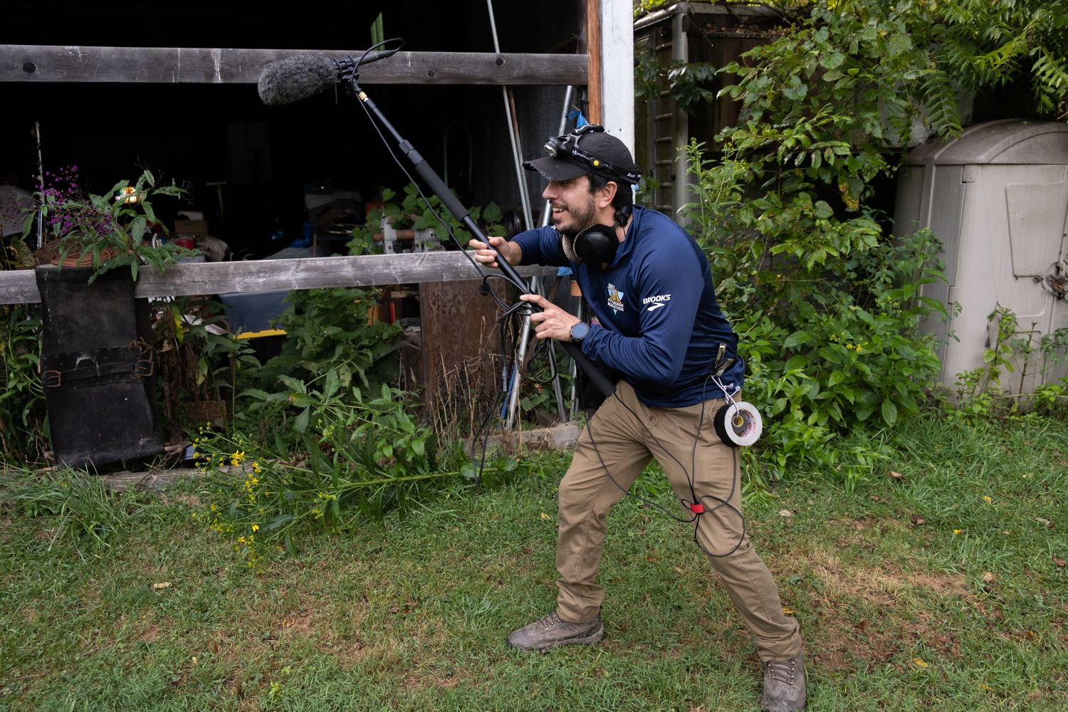 A boom operator holds a microphone pole while recording audio outdoors near a rustic building.