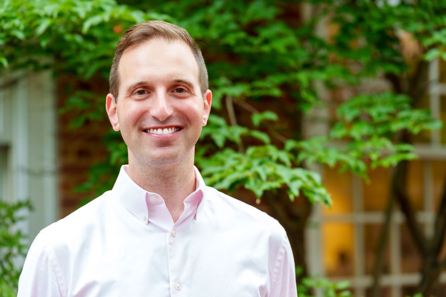 Smiling man in white shirt standing outdoors surrounded by lush green foliage in a garden setting.