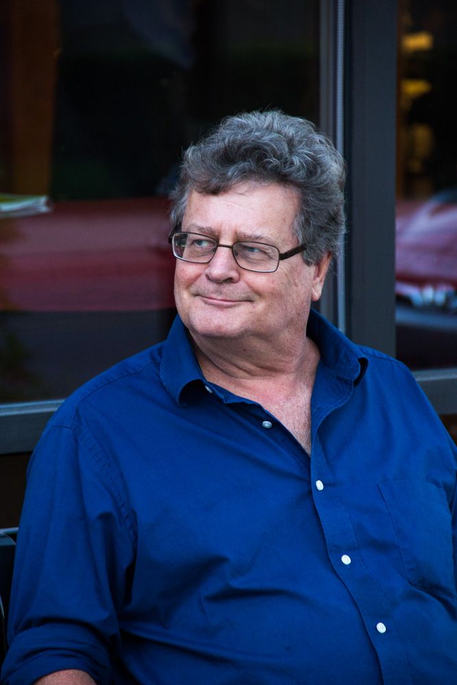 Older man with gray curly hair and glasses wearing a blue shirt, smiling outdoors near a glass window.