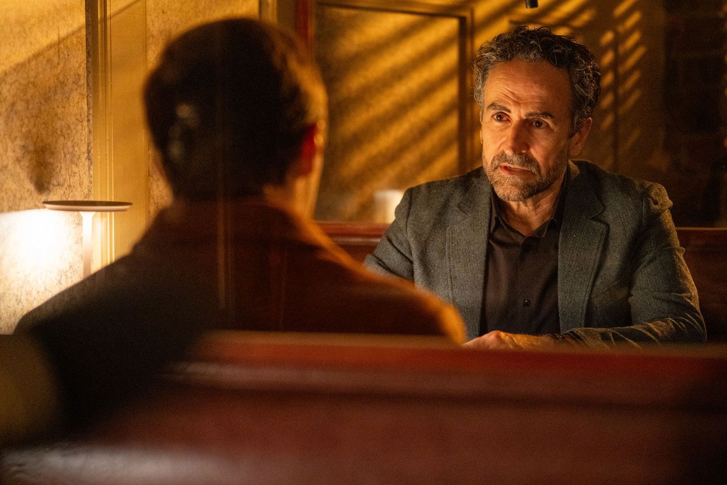 Two people in a dimly lit booth, man in gray blazer listens intently in warm, moody restaurant setting.
