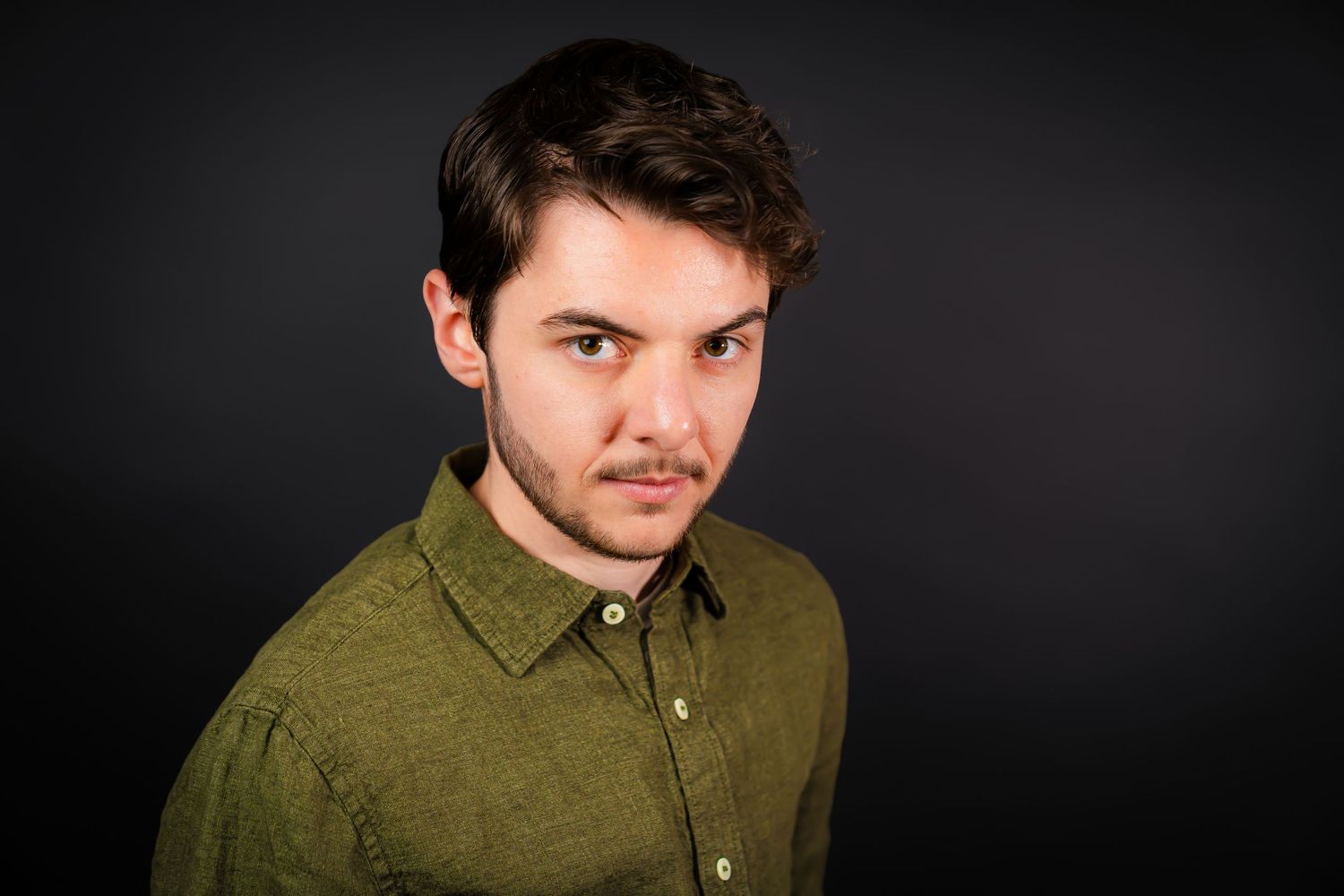 A man in an olive green button-up shirt poses against a dark background in a professional headshot.