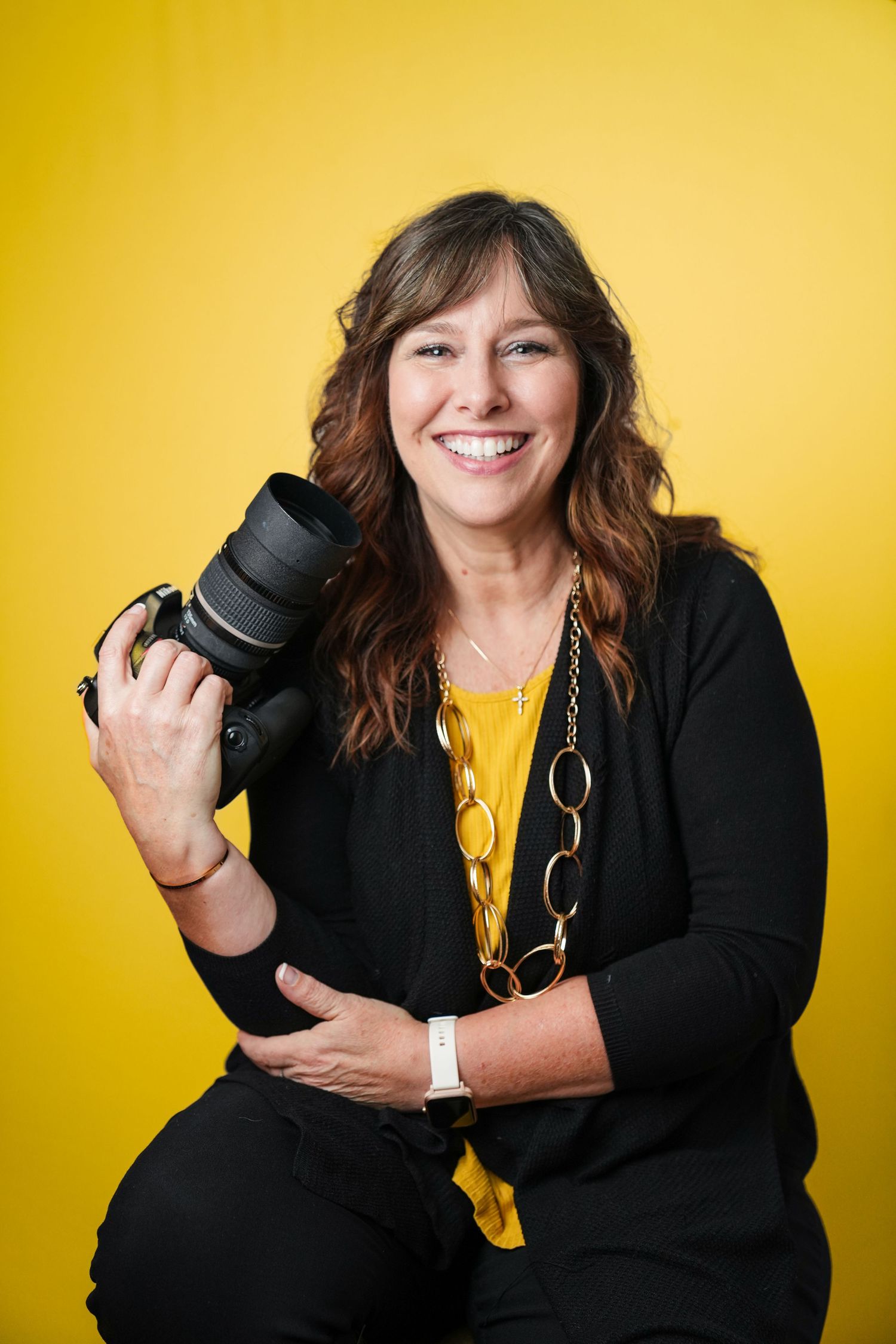 Professional photographer in black cardigan and yellow top holds DSLR camera against bright yellow background.