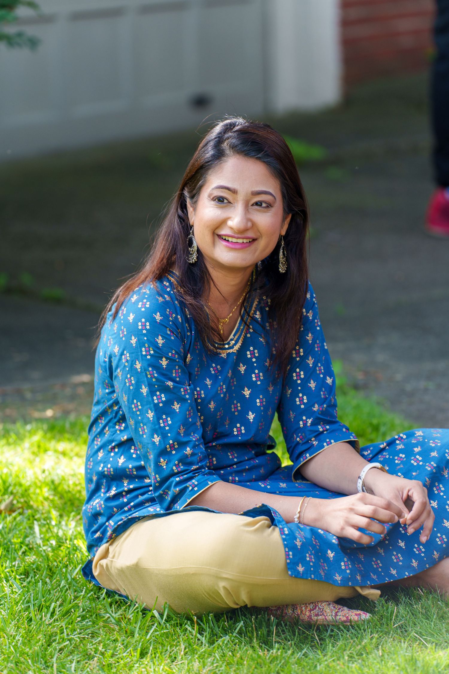 Smiling woman in blue floral kurta and gold earrings sitting cross-legged on green grass outdoors.
