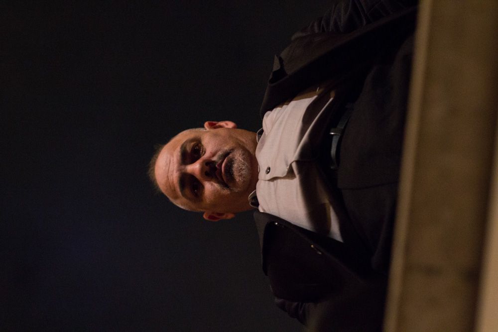 Bald man with beard in dark suit smiling, photographed in low-light setting against dark background.