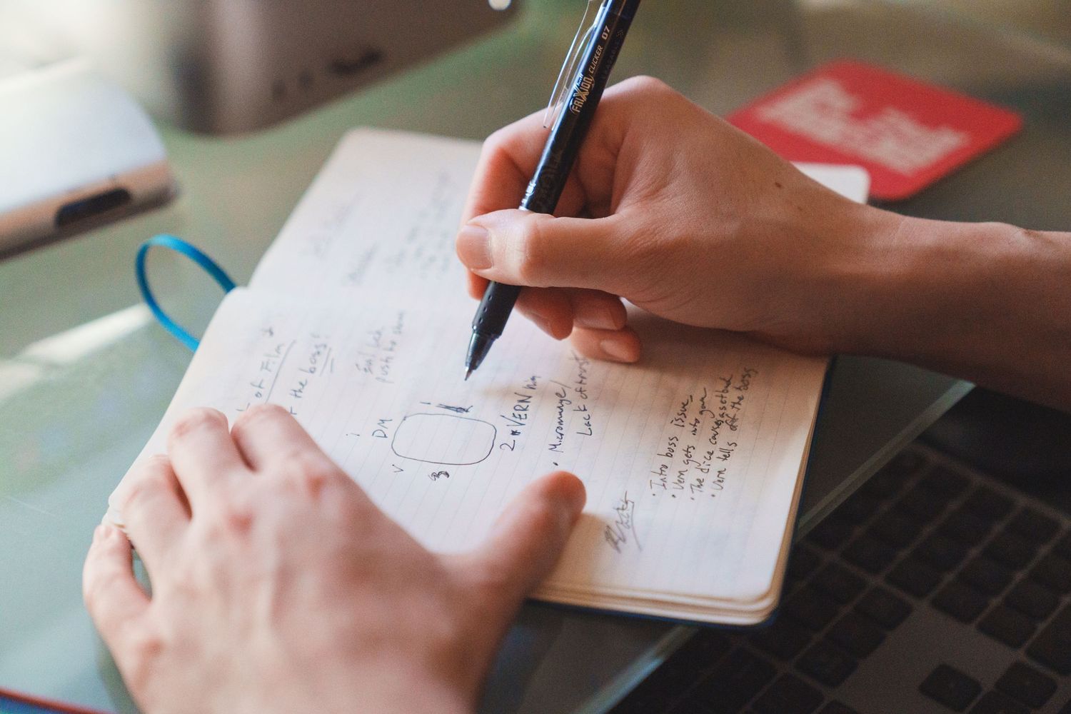 Close-up of hands writing notes in a notebook next to a laptop.