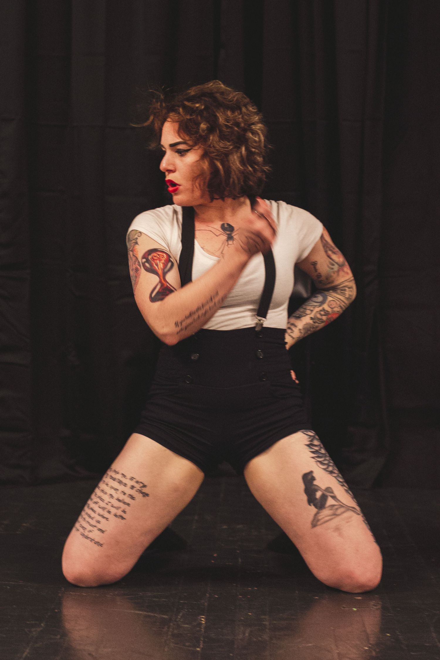 A figure in black performs an artistic dance pose on a dark stage with visible tattoos.