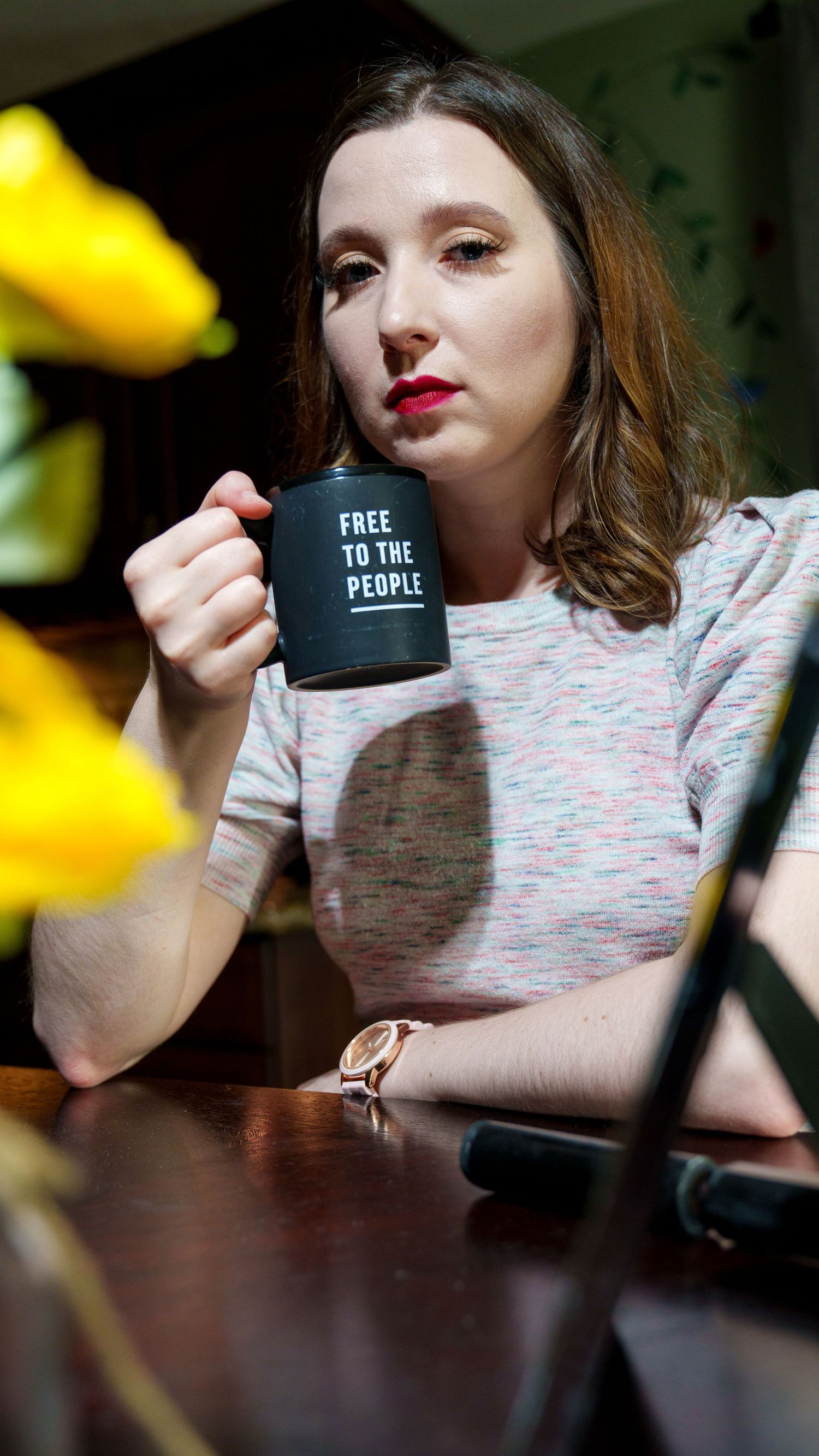 Woman sitting at table holding a black 'Free to the People' mug near yellow flowers, wearing a pink top and rose gold watch.