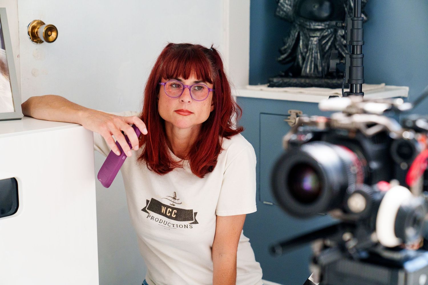 Red-haired woman in white t-shirt sits near camera on indoor film set with statue in background.