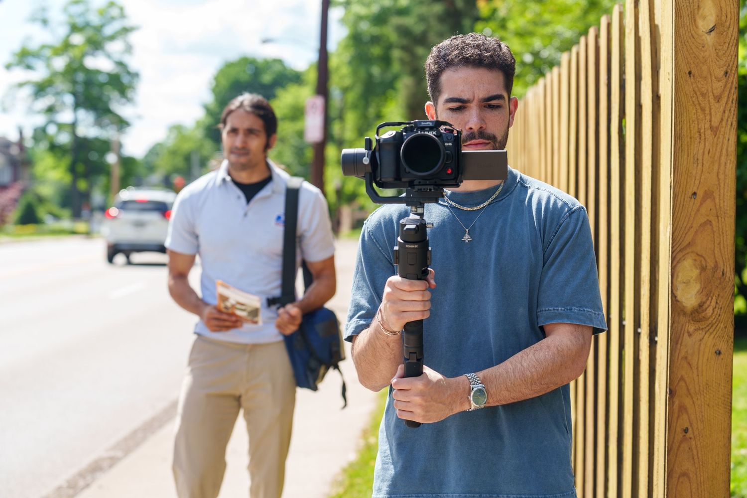 Two men filming outdoors near a wooden fence, one holding a camera on a gimbal stabilizer on a sunny day.