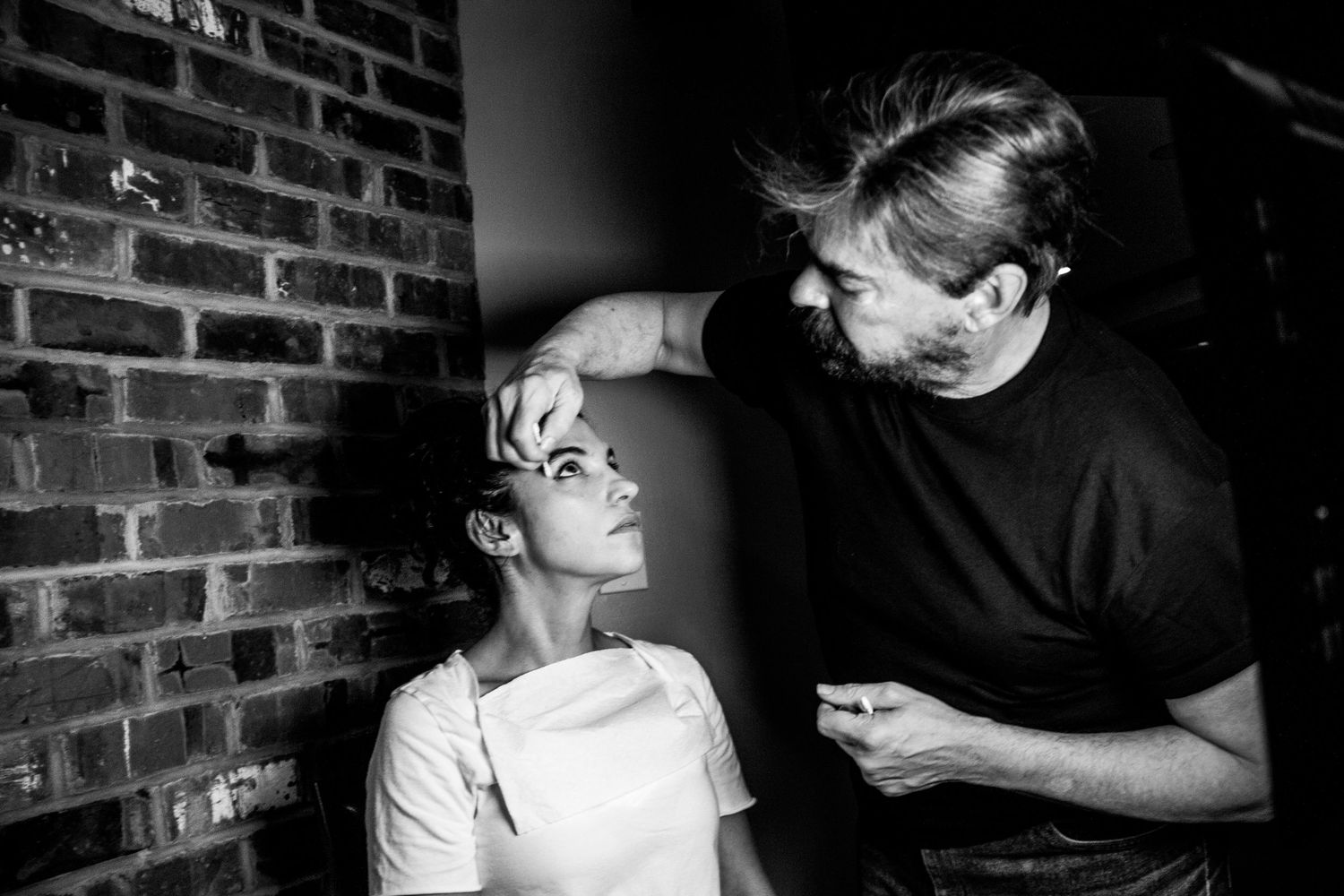 Black and white photo of a makeup artist applying makeup to a woman near a brick wall.