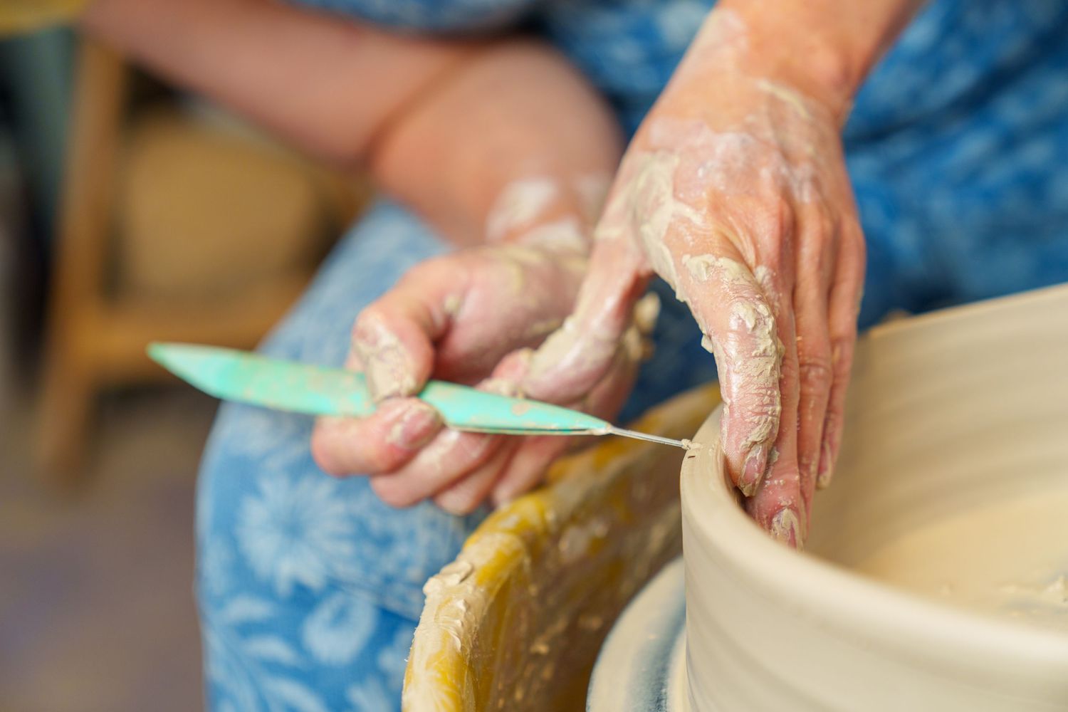 Hands covered in clay use a pottery tool to shape and smooth the rim of a ceramic vessel.