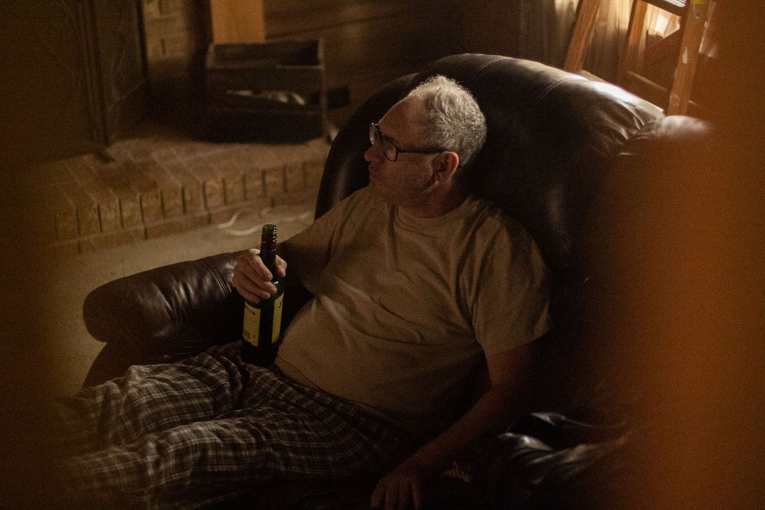 Older man relaxing in leather armchair holding a bottle in dimly lit room with warm ambient lighting.