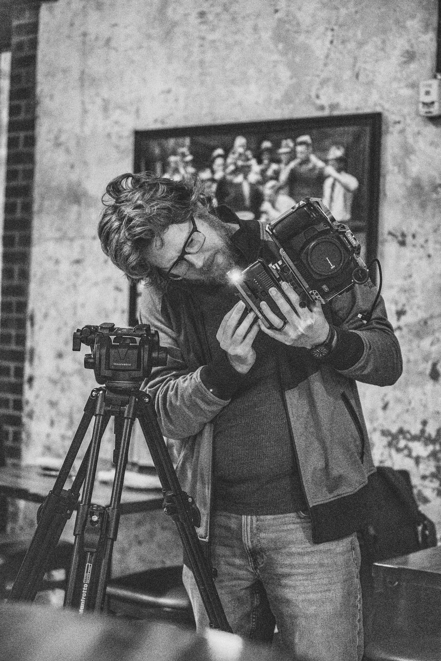 Bearded man in glasses holding a medium format camera next to a tripod in a black and white photo.
