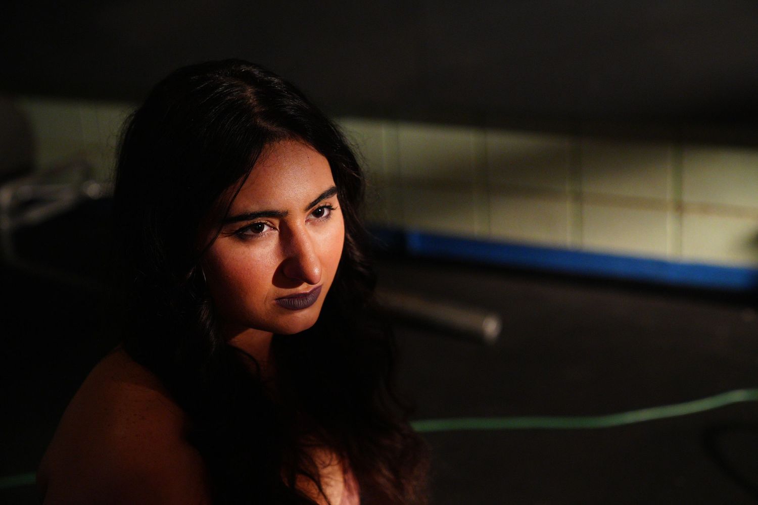 A young woman with long dark hair poses in dramatic lighting near a boxing ring.