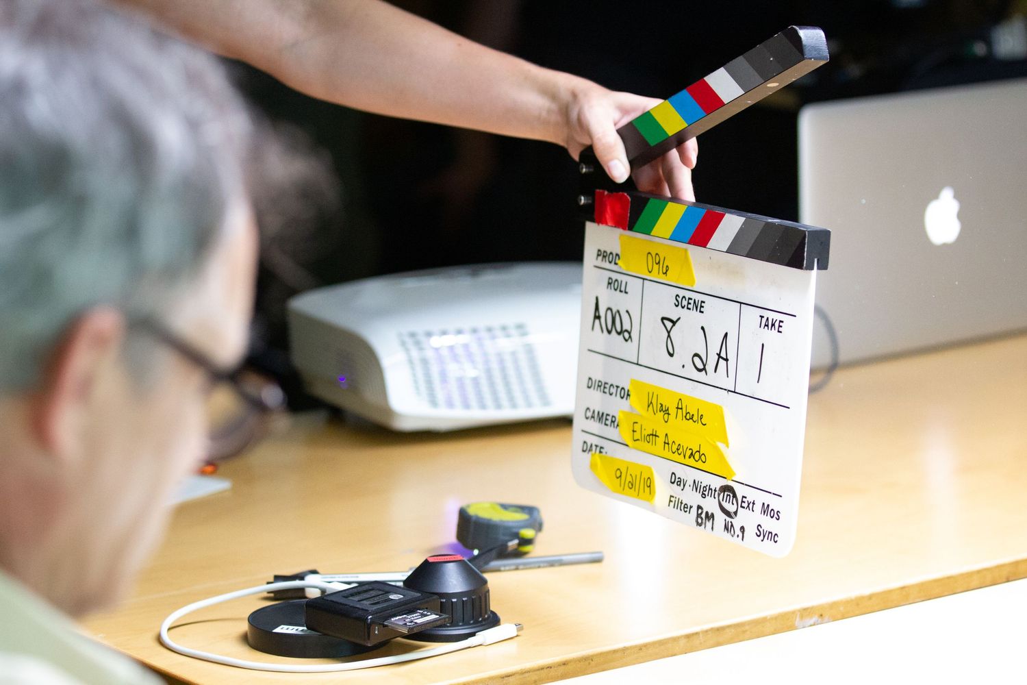 A film crew member holds a clapperboard on a movie set with an Apple laptop visible in the background.