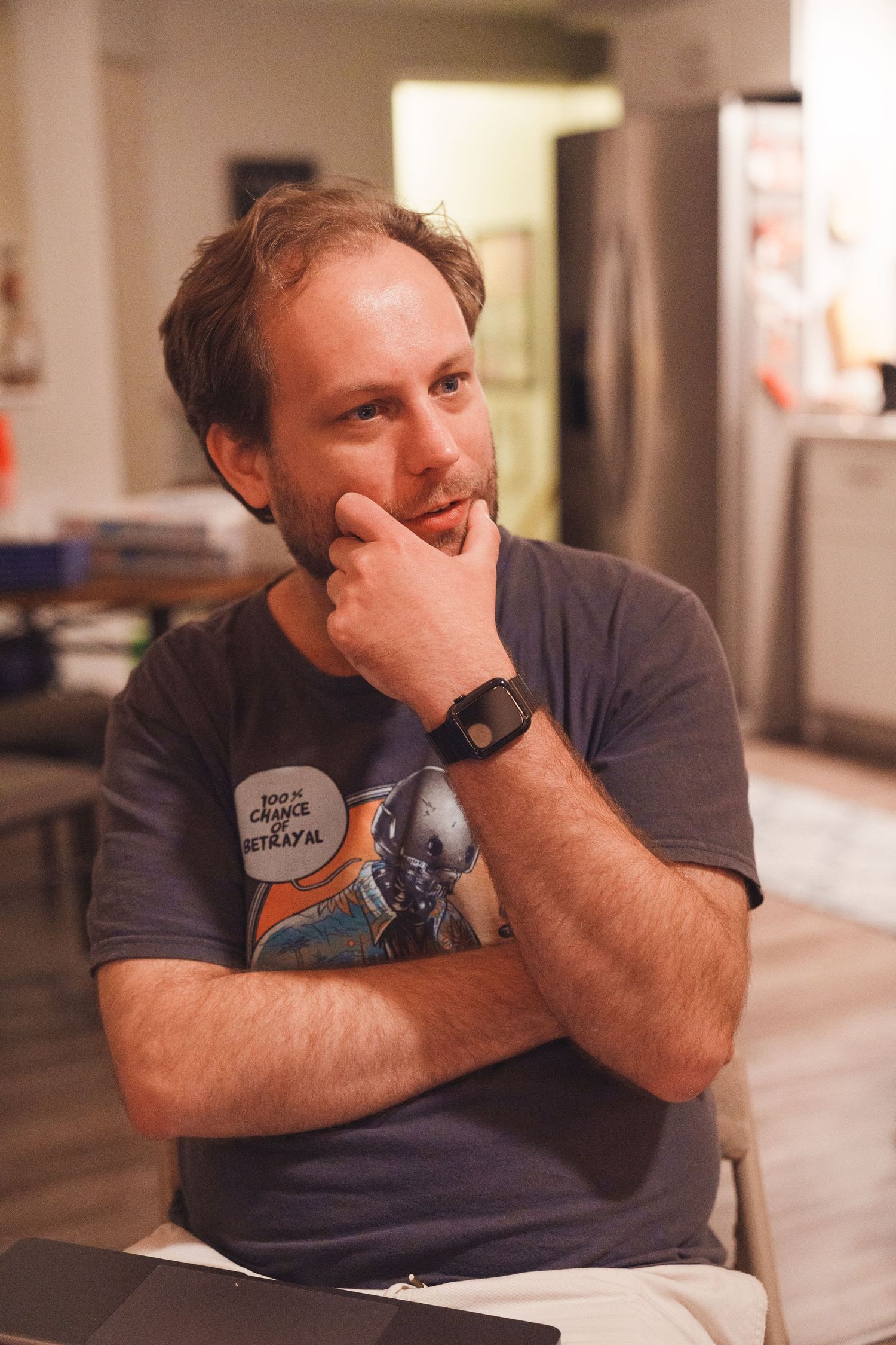 Thoughtful man with stubble wearing graphic tee sits with hand on chin in casual home setting.