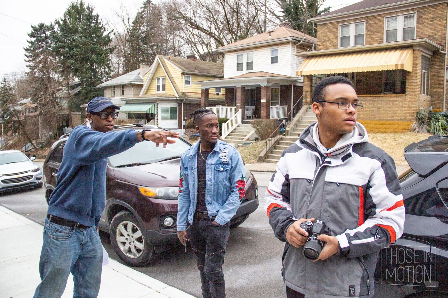 Three young men on a residential street, one pointing directions, another holding a camera, in an urban neighborhood.