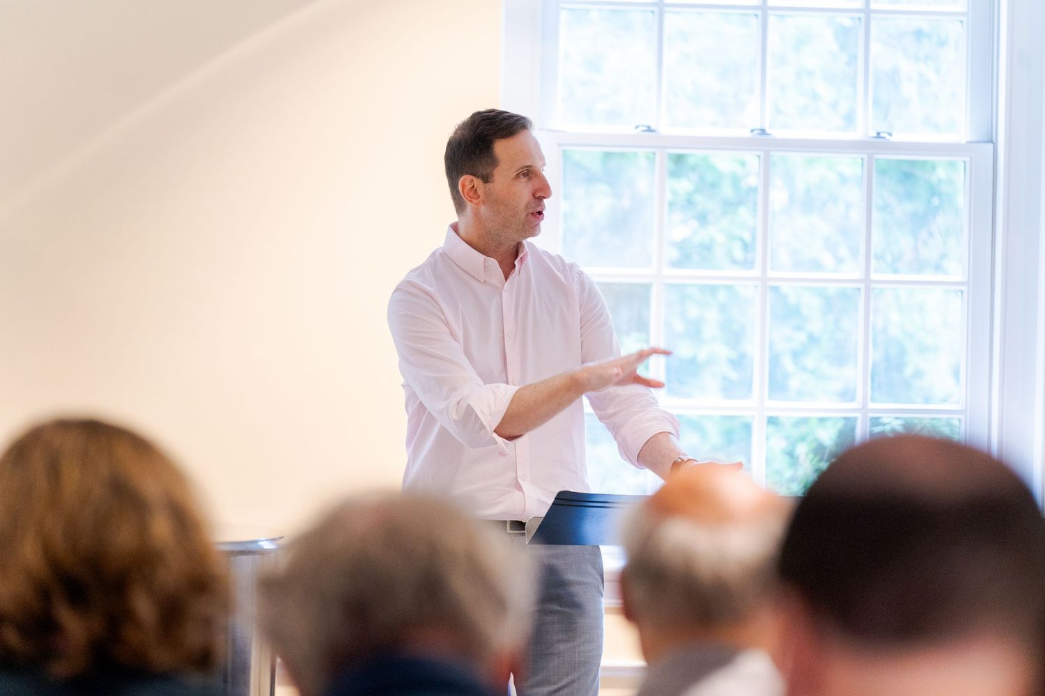 Man in white shirt gestures while presenting to an audience in a bright, sunlit room.