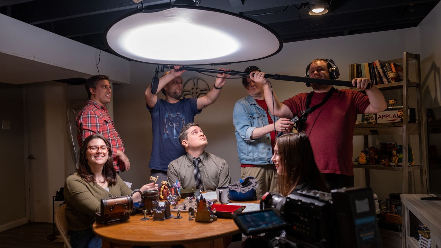 Six people pose playfully in a basement podcast studio with professional lighting, microphones, and recording equipment.