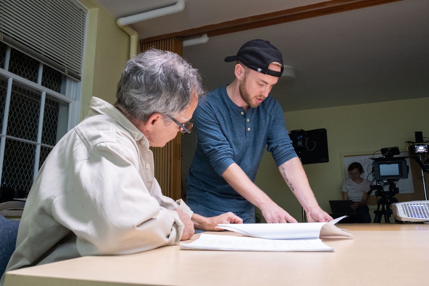 Two men review documents on a table in a production office with a camera visible in the background.