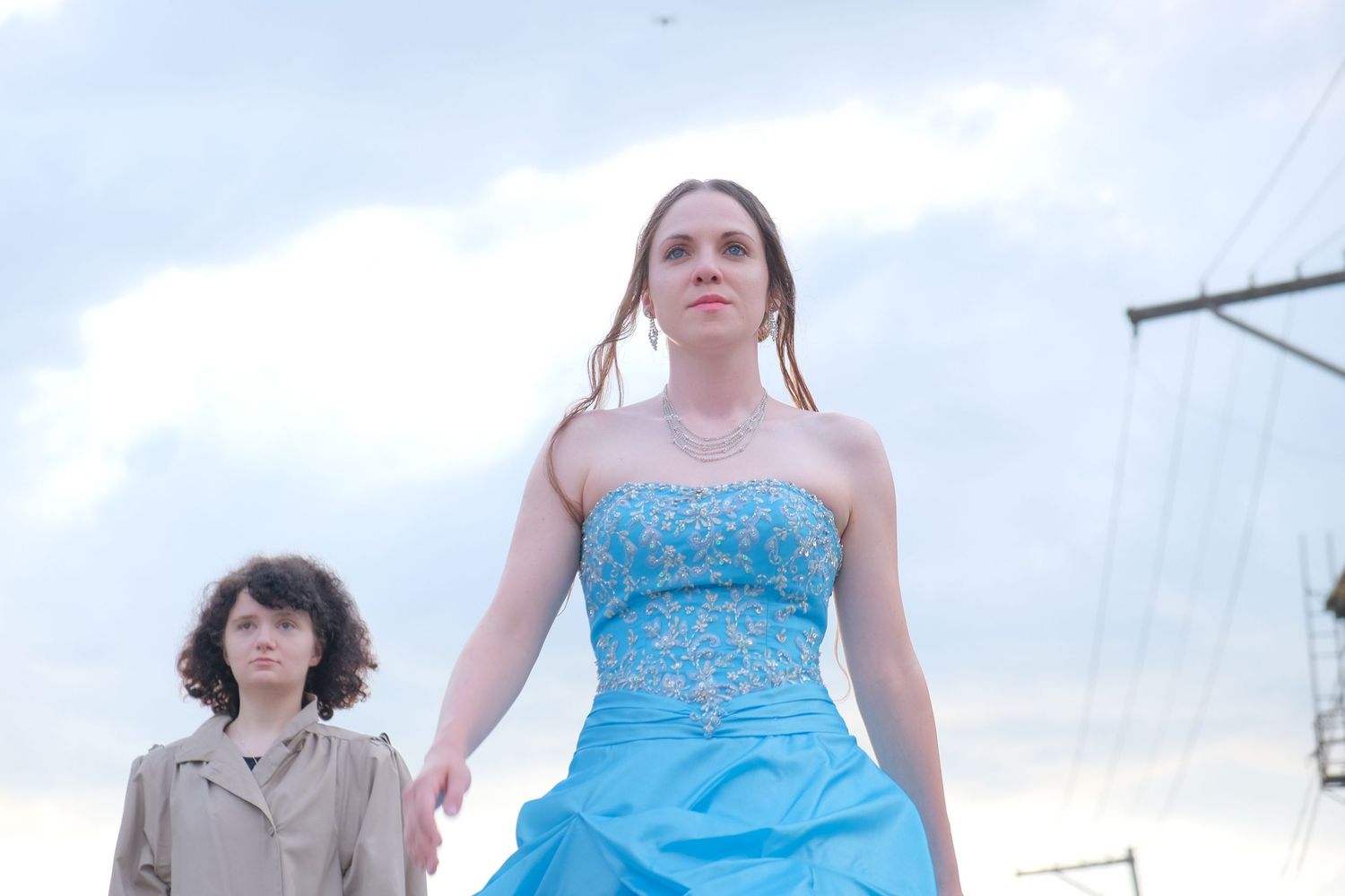 Two women outdoors, one in a blue strapless gown, posing against a cloudy sky near power lines.