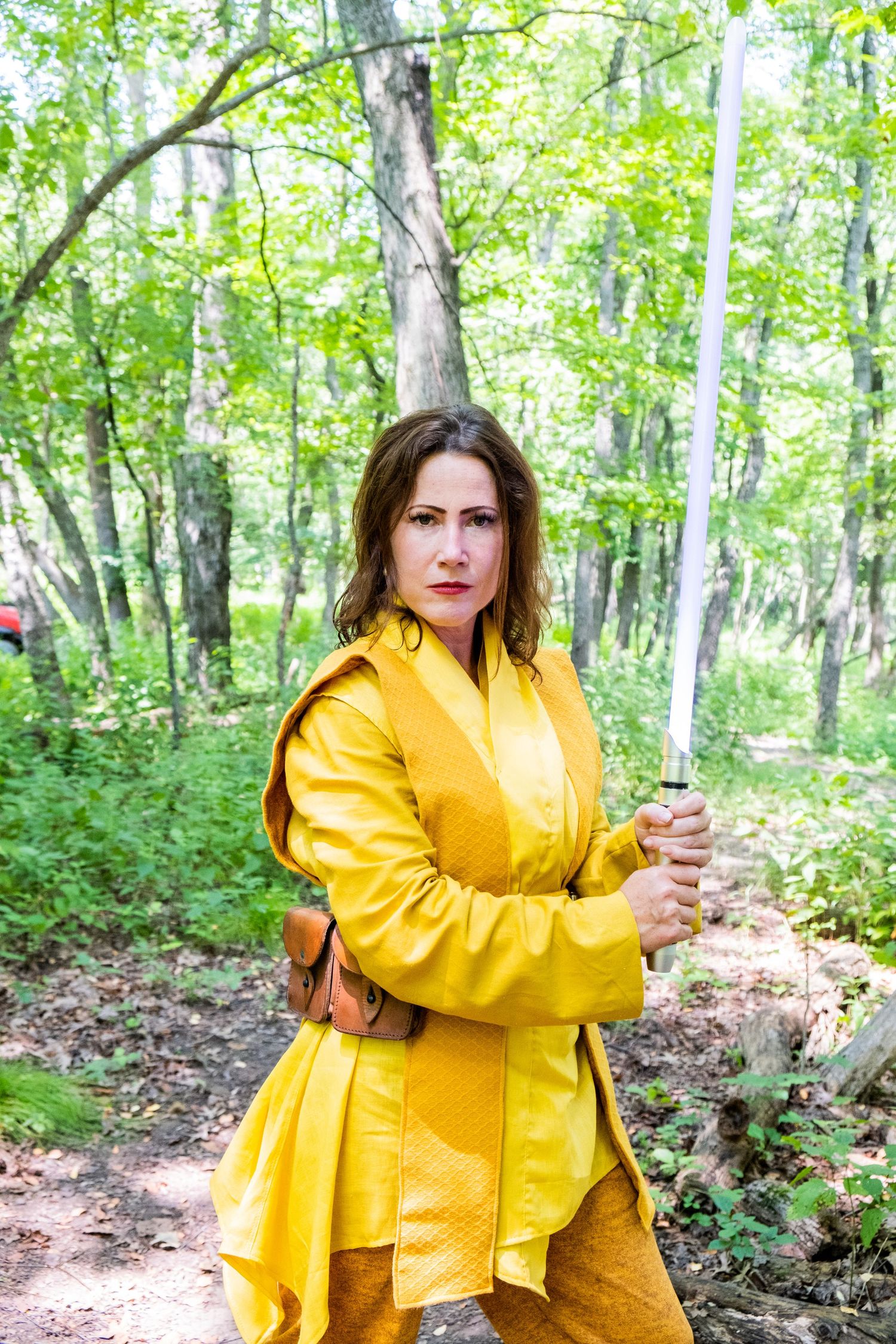 A figure in a yellow raincoat poses with a lightsaber in a bright forest setting.