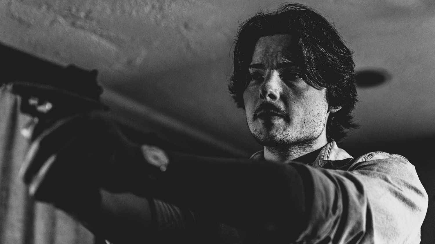 Black and white film noir style shot of person aiming weapon in dramatic lighting.
