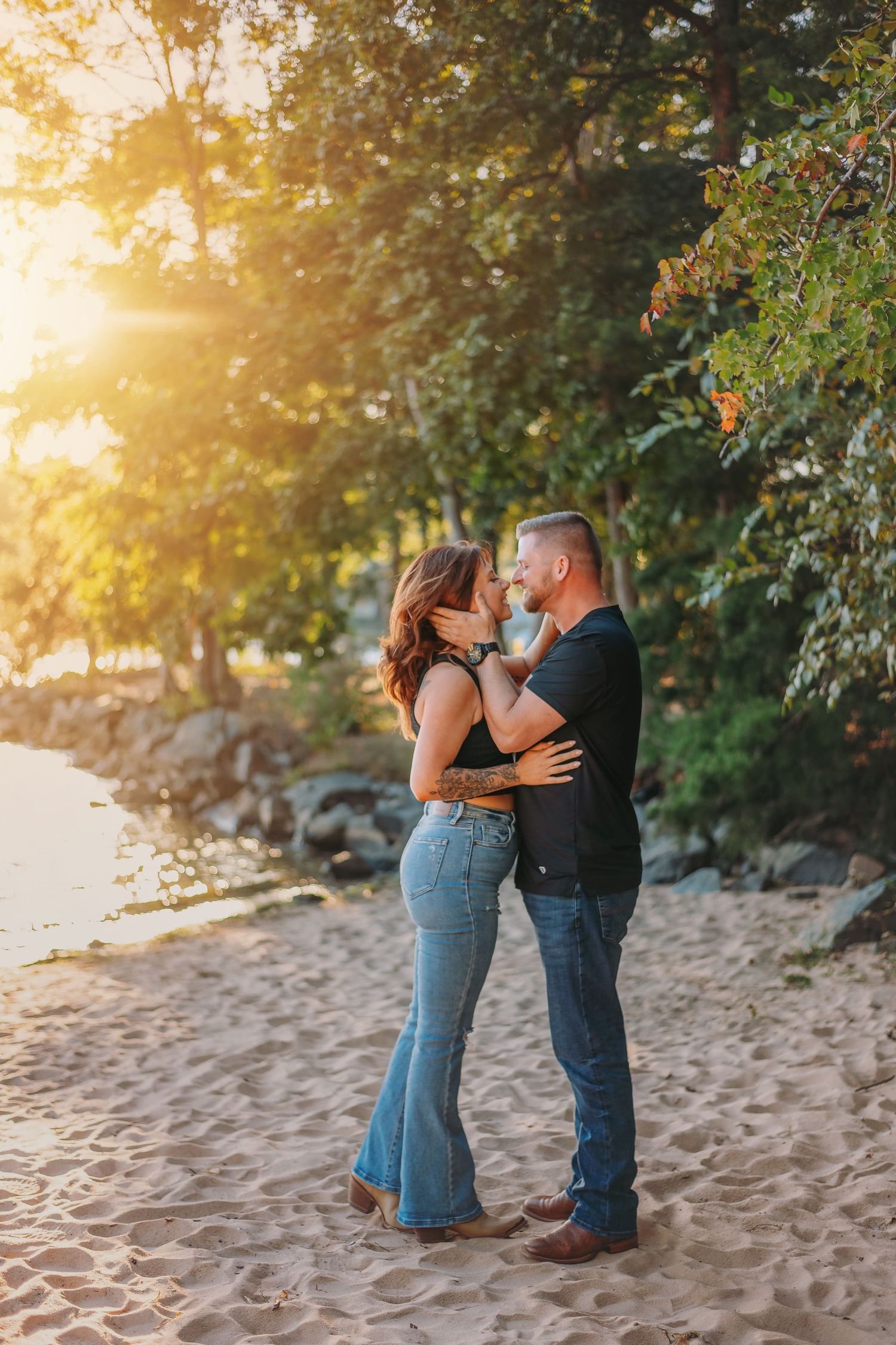 Couple embracing on sandy beach during golden hour with trees in background.