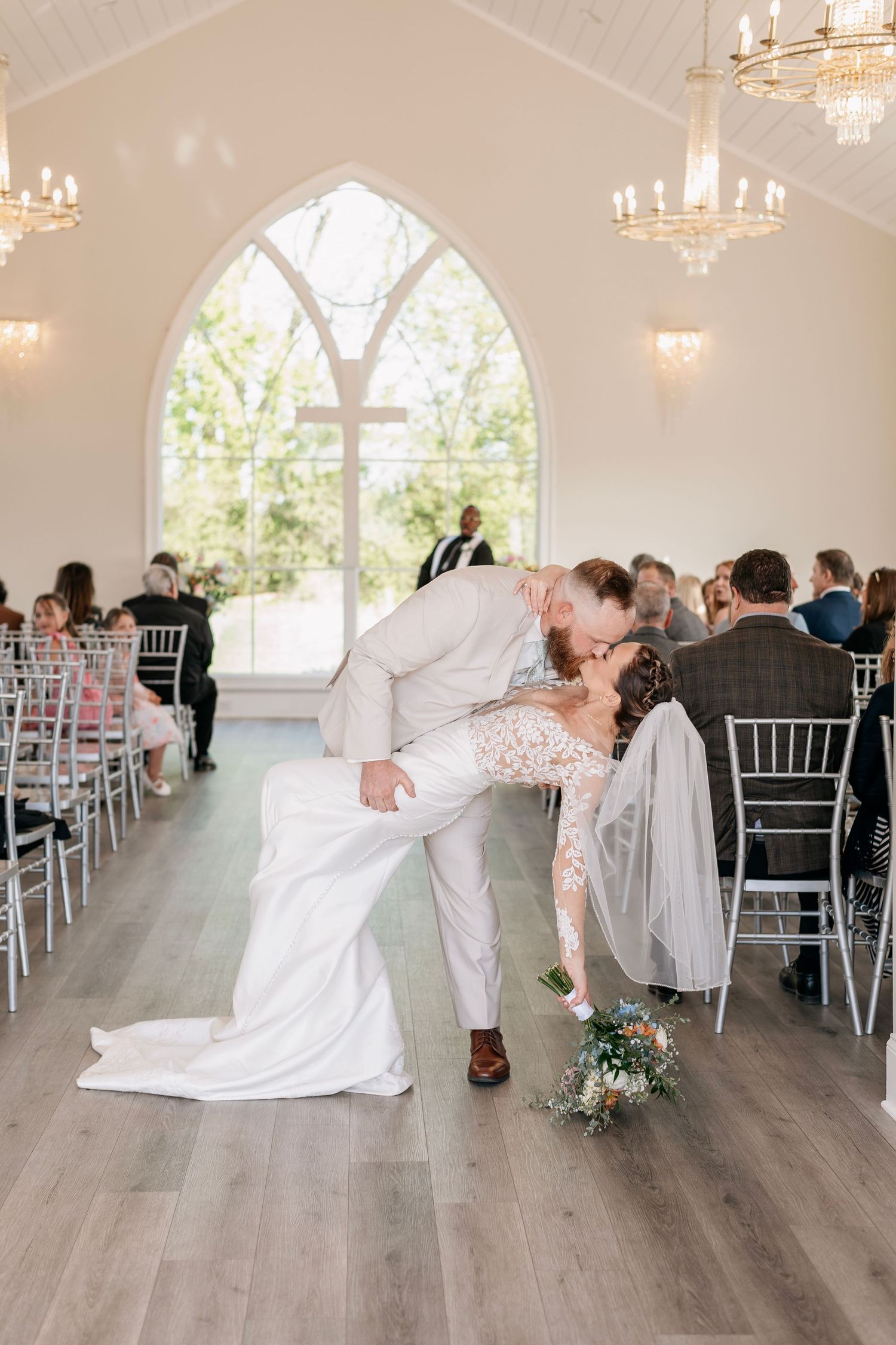 Bride and groom share a romantic dip kiss in a bright white wedding chapel aisle with chandeliers and seated guests.