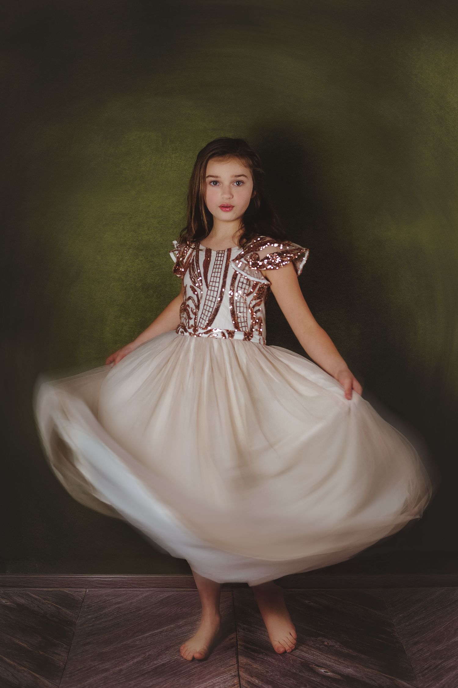 A young child in a flowing cream dress twirls and dances barefoot against a dark moody backdrop.