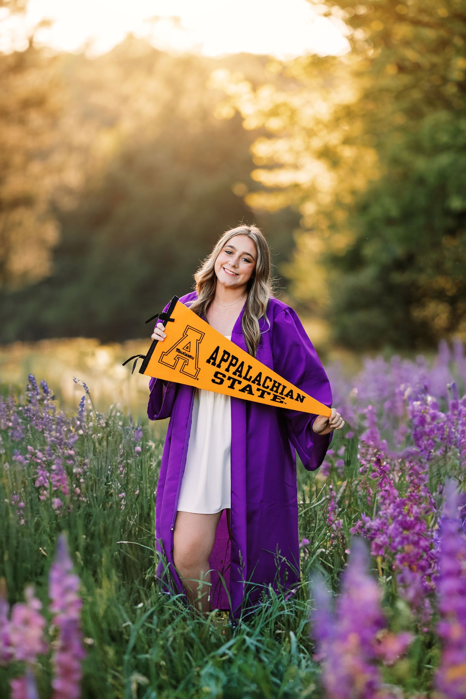 Graduate in purple gown holding Appalachian pennant among purple wildflowers at sunset.
