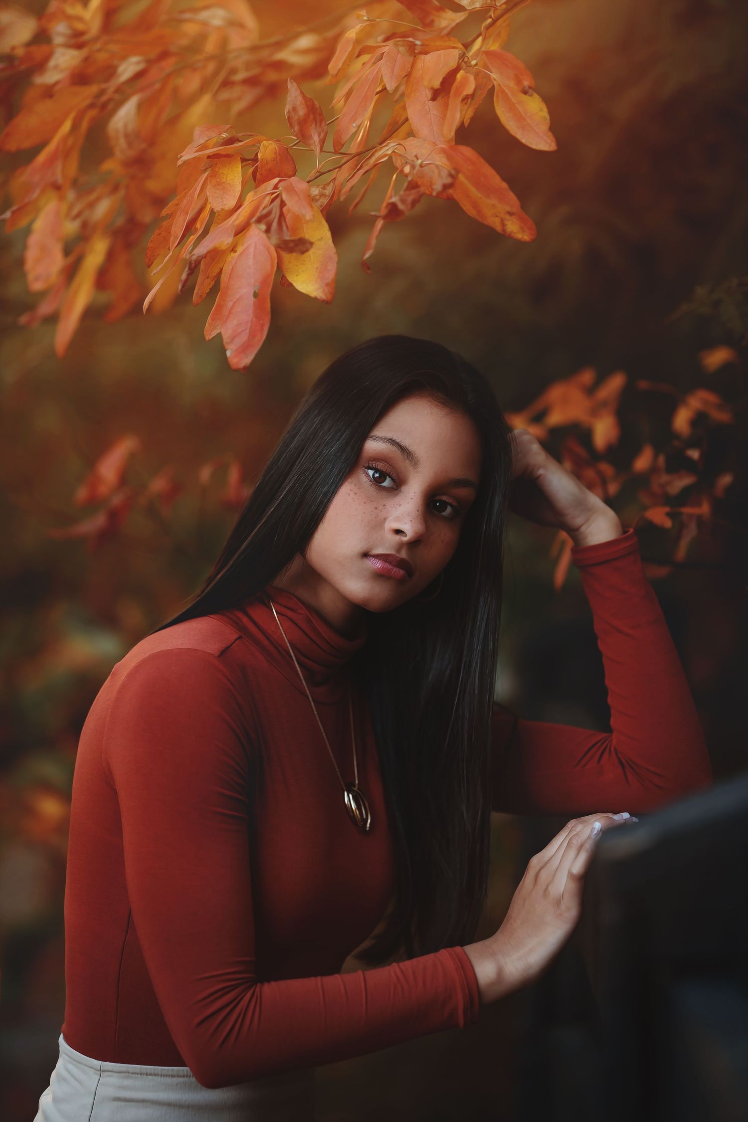 Woman in red turtleneck poses under autumn leaves in moody lighting.